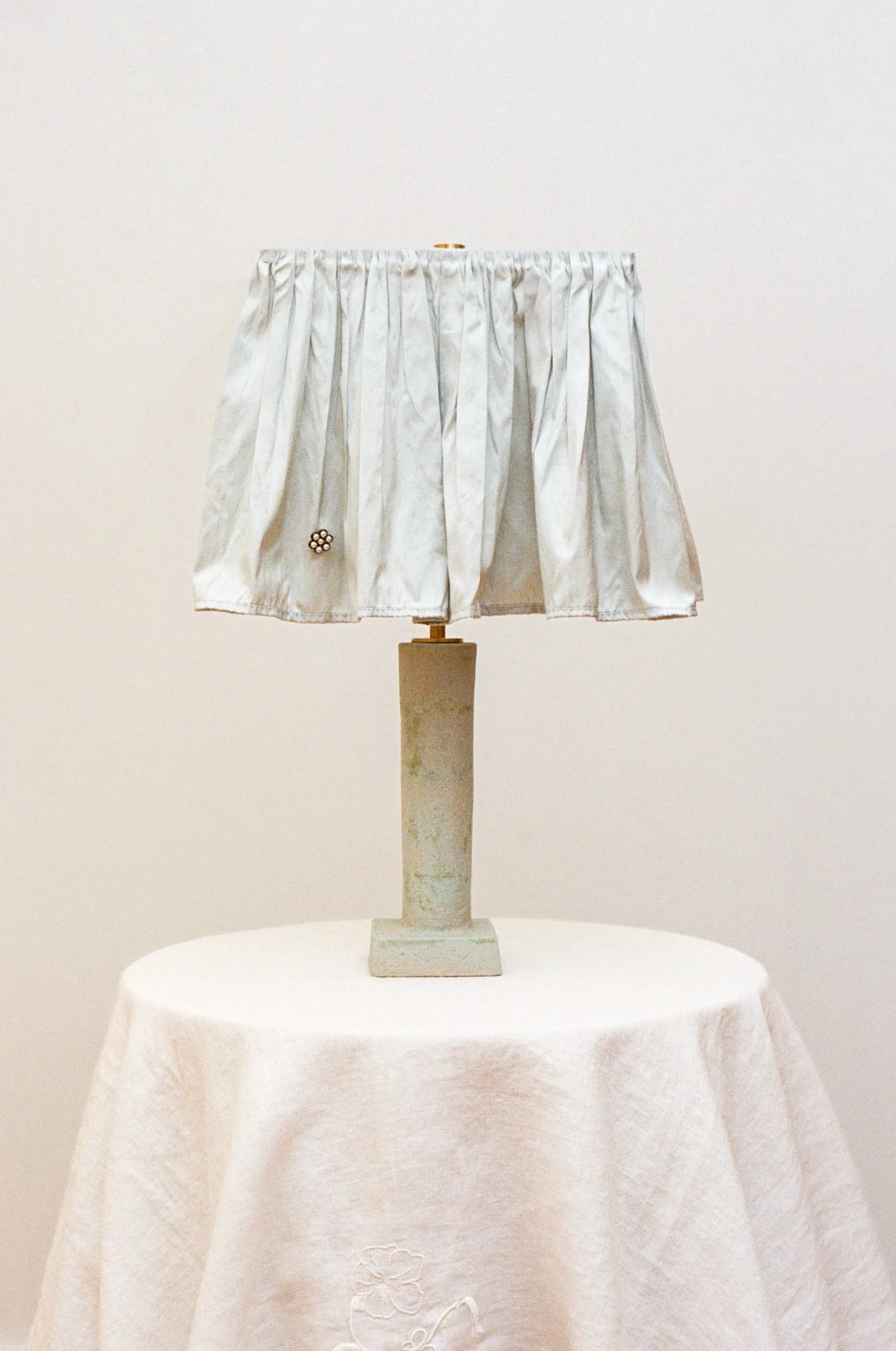 A table lamp with a weathered brass cylindrical base and pleated linen shade in cream, positioned on a round white tablecloth by Analuisa Corrigan.