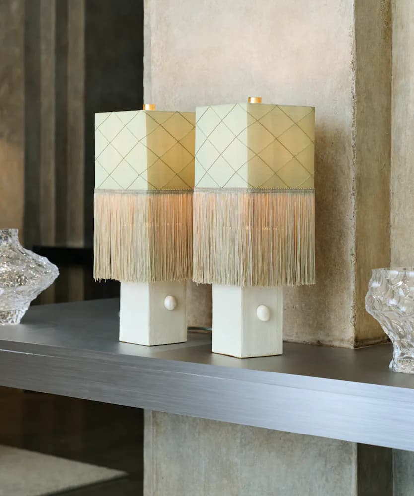 Two white cubic table lamps with quilted cream and gold shades topped with natural fringe trim, displayed on a gray shelf against textured walls by Analuisa Corrigan.