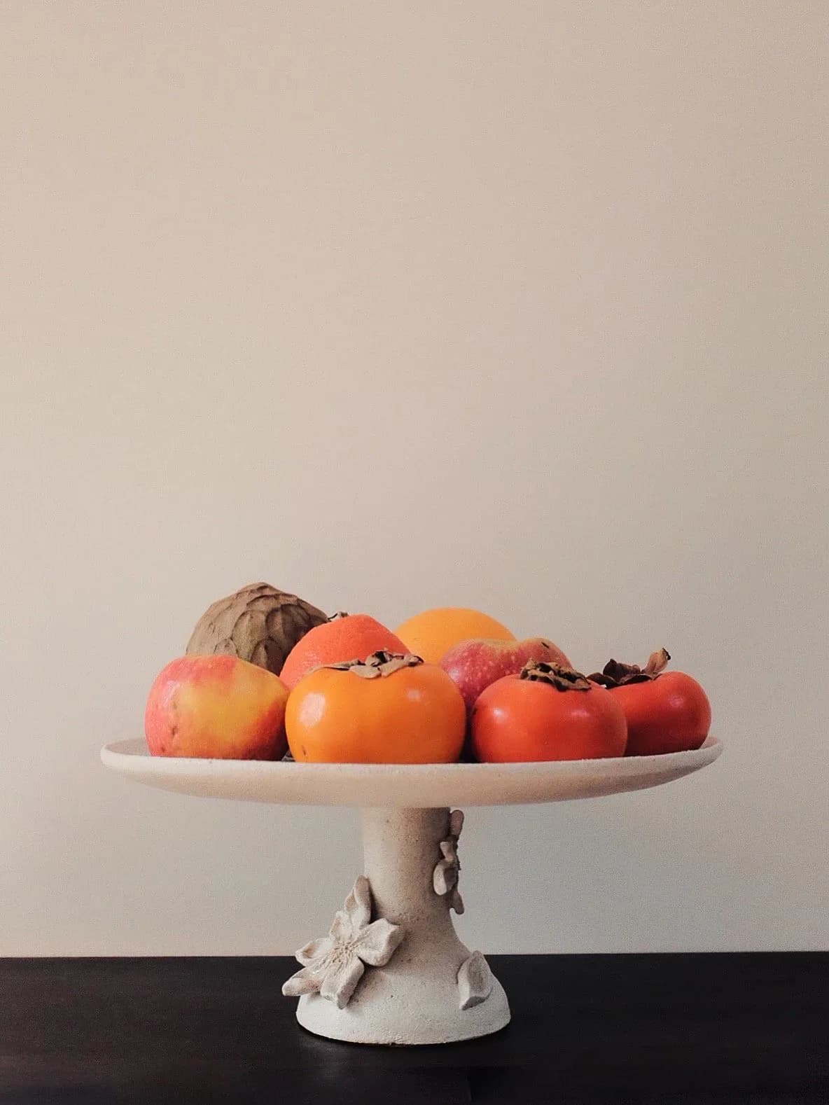 Styled still life arrangement of fresh fruits on a decorative pedestal bowl against a neutral background, by Analuisa Corrigan.