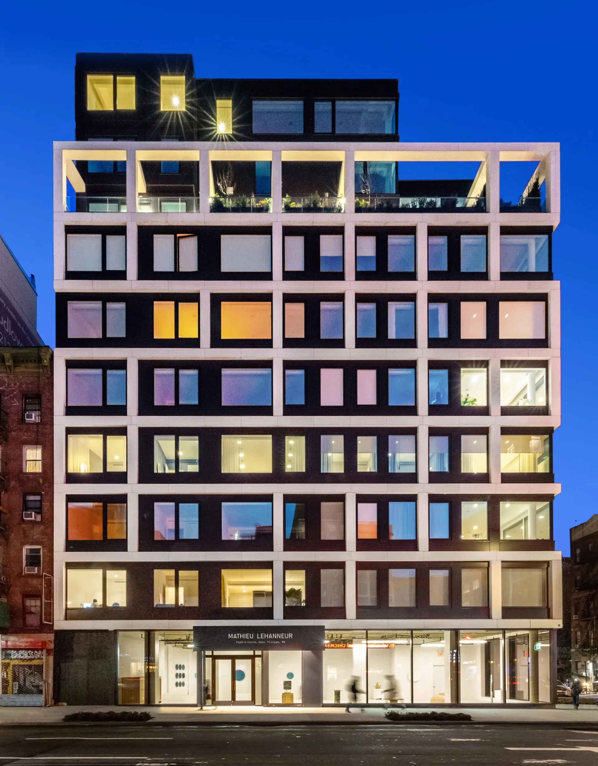 Modern multi-story residential building with illuminated windows in varied colors against evening sky, featuring geometric grid facade design by Anam Ahmed.