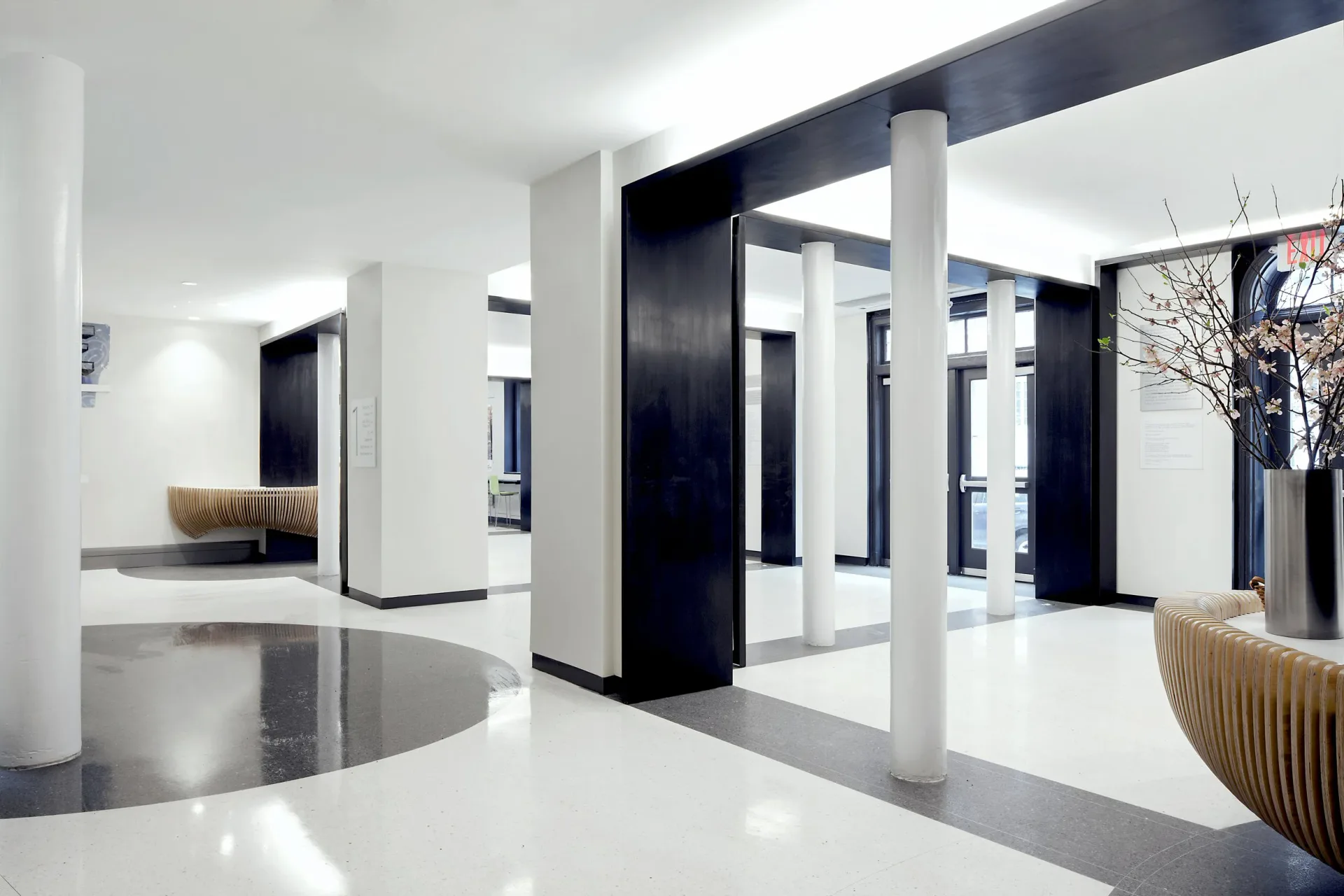 Minimalist interior space with white walls, black structural elements, polished concrete floors, and cylindrical sculptural forms by Anam Ahmed.