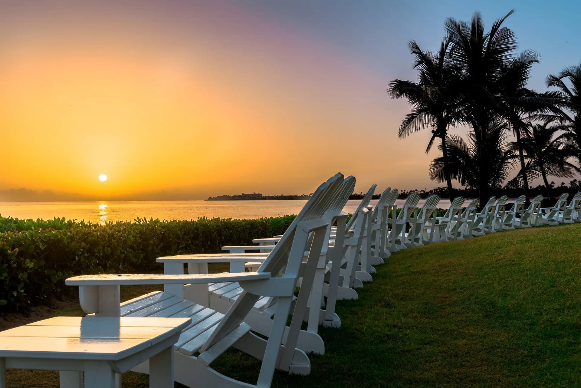 Waterfront seating area at sunset with white Adirondack chairs overlooking water and palm trees, designed by Andrea Rodriguez.