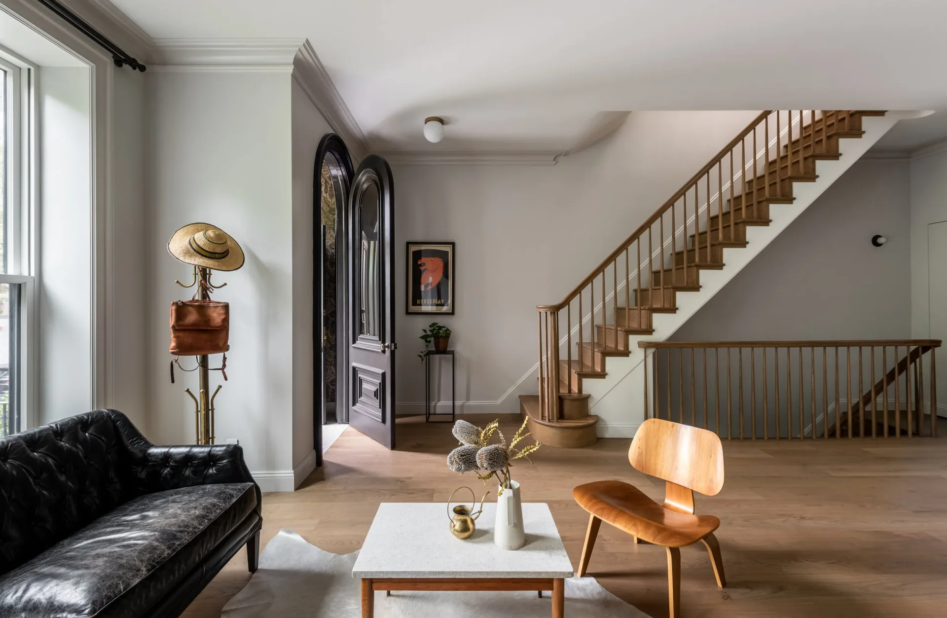A modern living room features a black sofa, wooden furniture, and a central staircase with metal railings; light wood flooring and neutral tones throughout by Andrew Bernheimer.