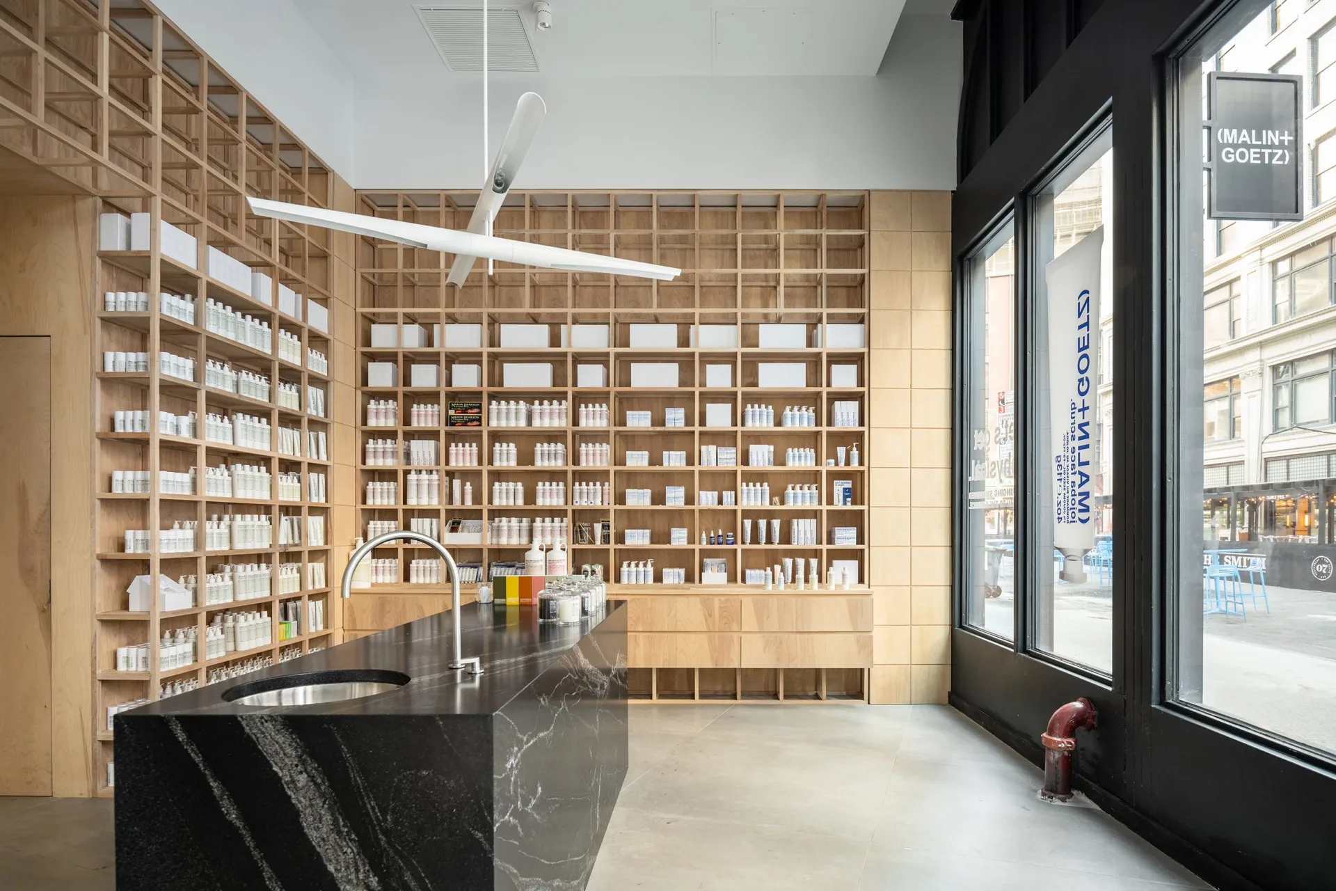 Modern pharmacy interior with floor-to-ceiling wooden shelving, black marble counter, and large storefront windows overlooking a street by Andrew Bernheimer.