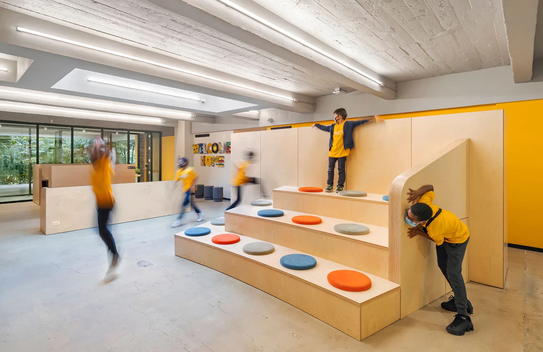 Bright educational interior with modular wooden stepped platforms in natural wood and colorful circular elements, featuring yellow accent walls and linear ceiling lights, by Andrew Franz.