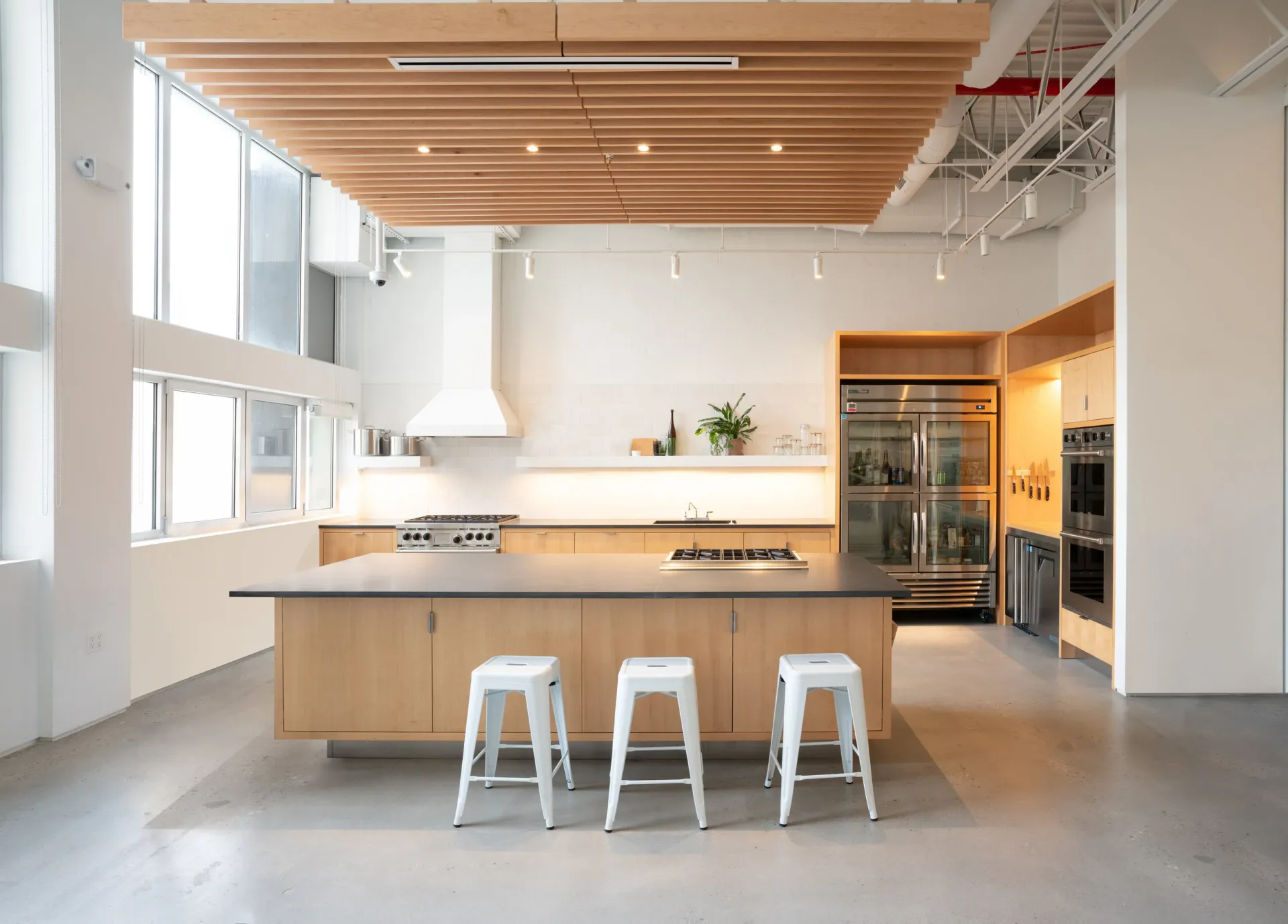 Modern kitchen with light wood cabinetry, white countertops, and a central island with white stools, polished concrete floors, and large windows by Andrew L. Pettit.
