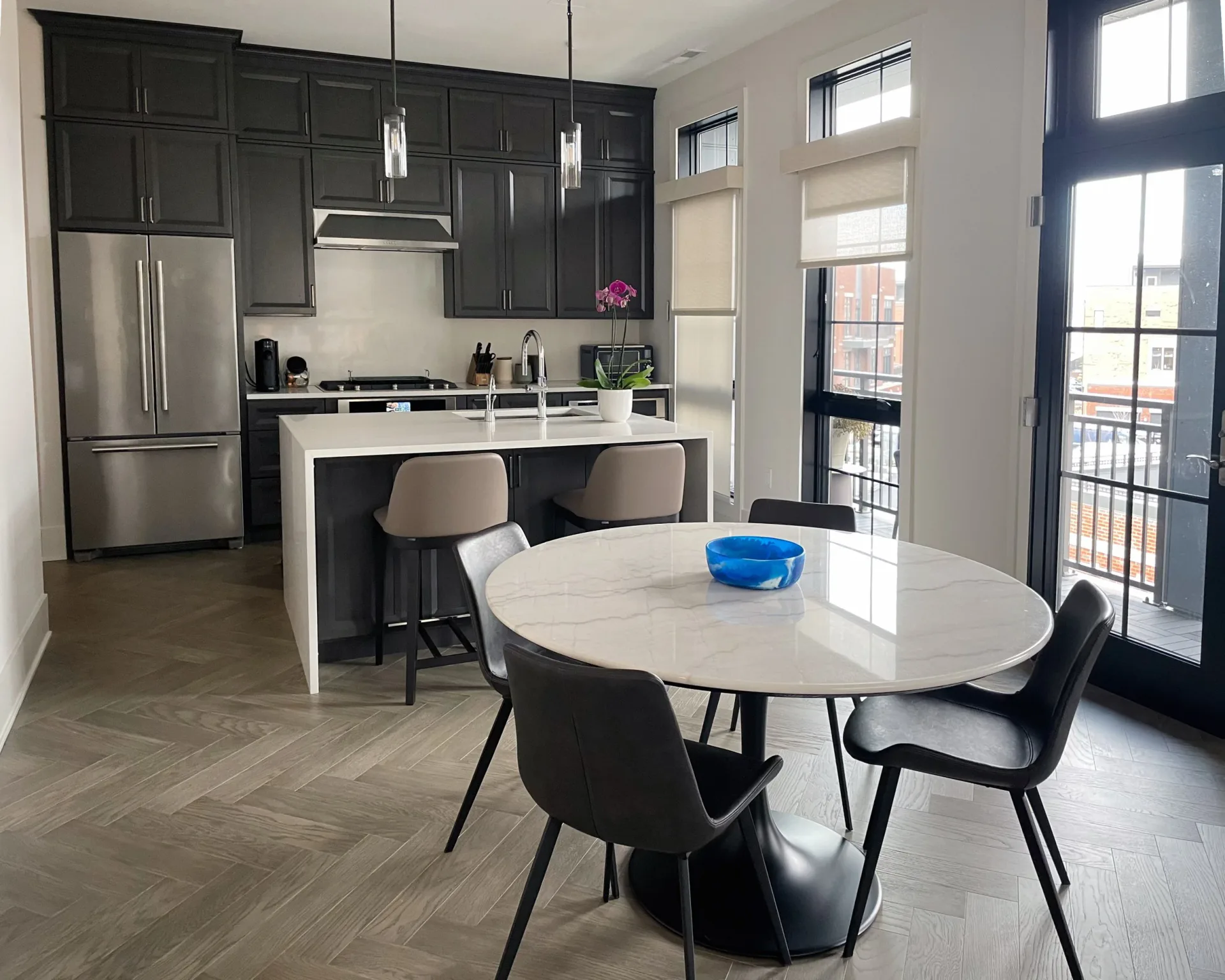 Modern open-concept kitchen and dining area with dark cabinetry, stainless steel appliances, white marble dining table, black chairs, and herringbone flooring by Andy Schwalen.