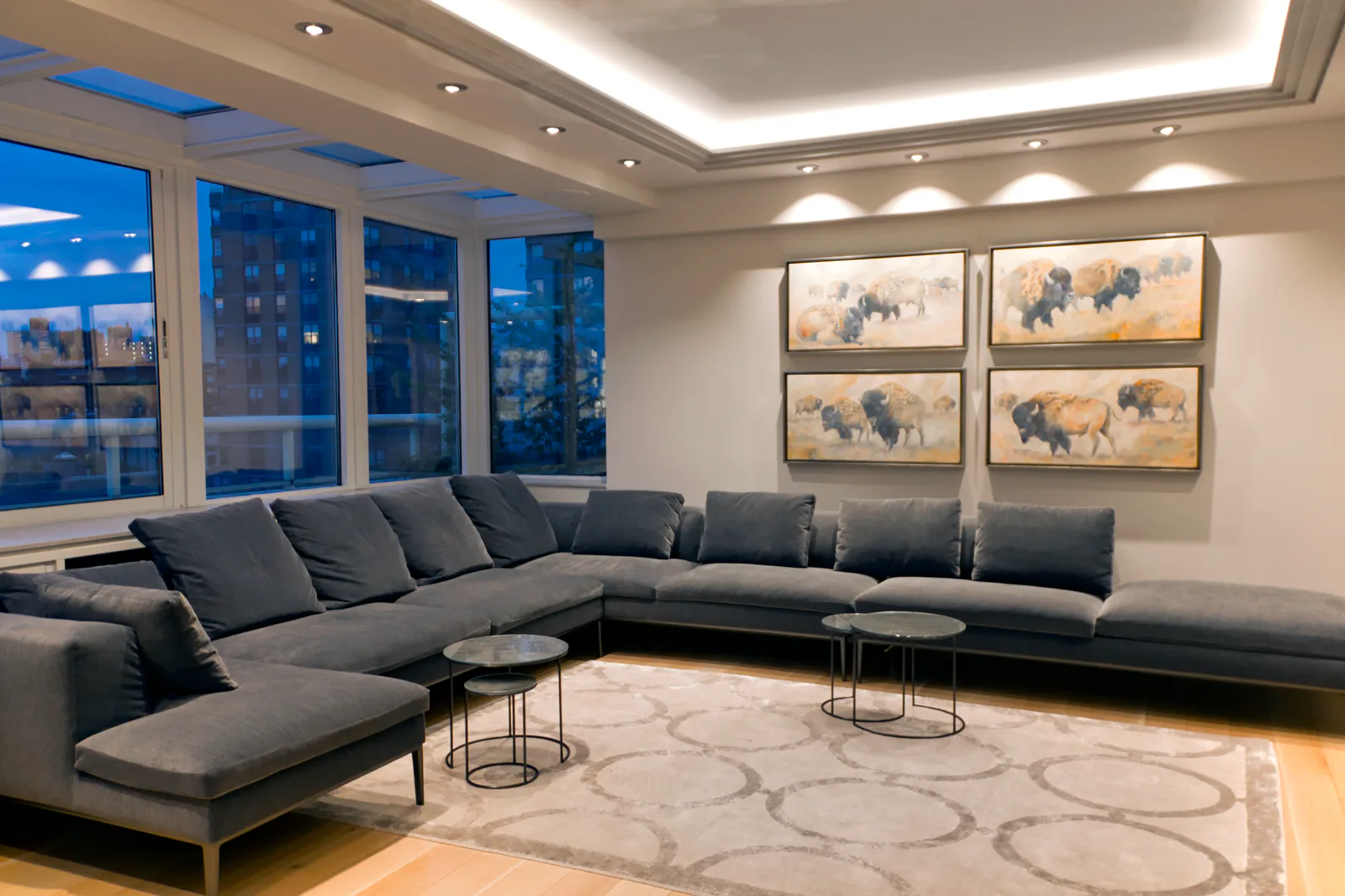 Modern living room with gray sectional sofa, marble coffee tables, large windows with city views, elephant artwork, and ambient ceiling lighting by Andy Schwalen.