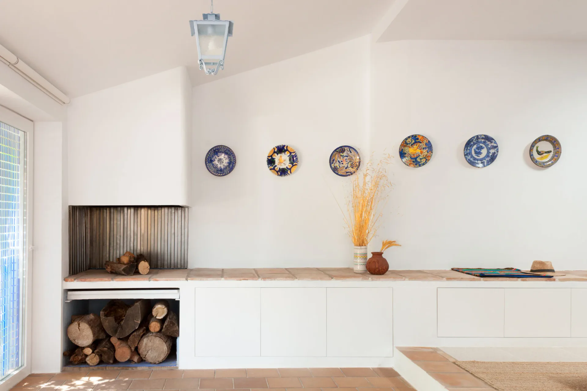 Minimalist kitchen with white cabinetry, wooden countertops, decorative blue plates on wall, and firewood storage, designed by Angel Mombiedro (Armombiedro Studio).