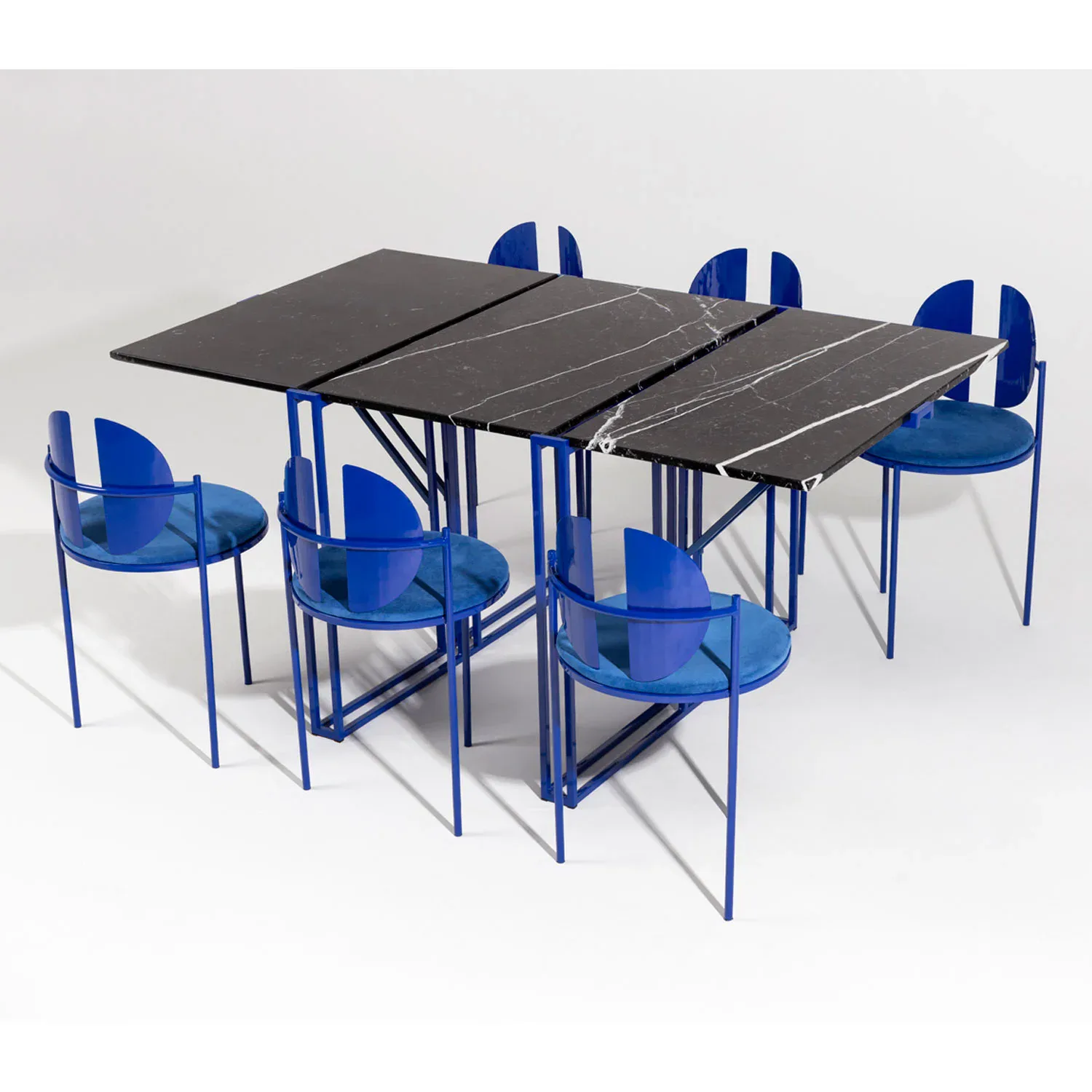 Dining set with extendable black table and six blue transparent chairs arranged around it, featuring minimalist metal frame construction by Angel Mombiedro (Armombiedro Studio).