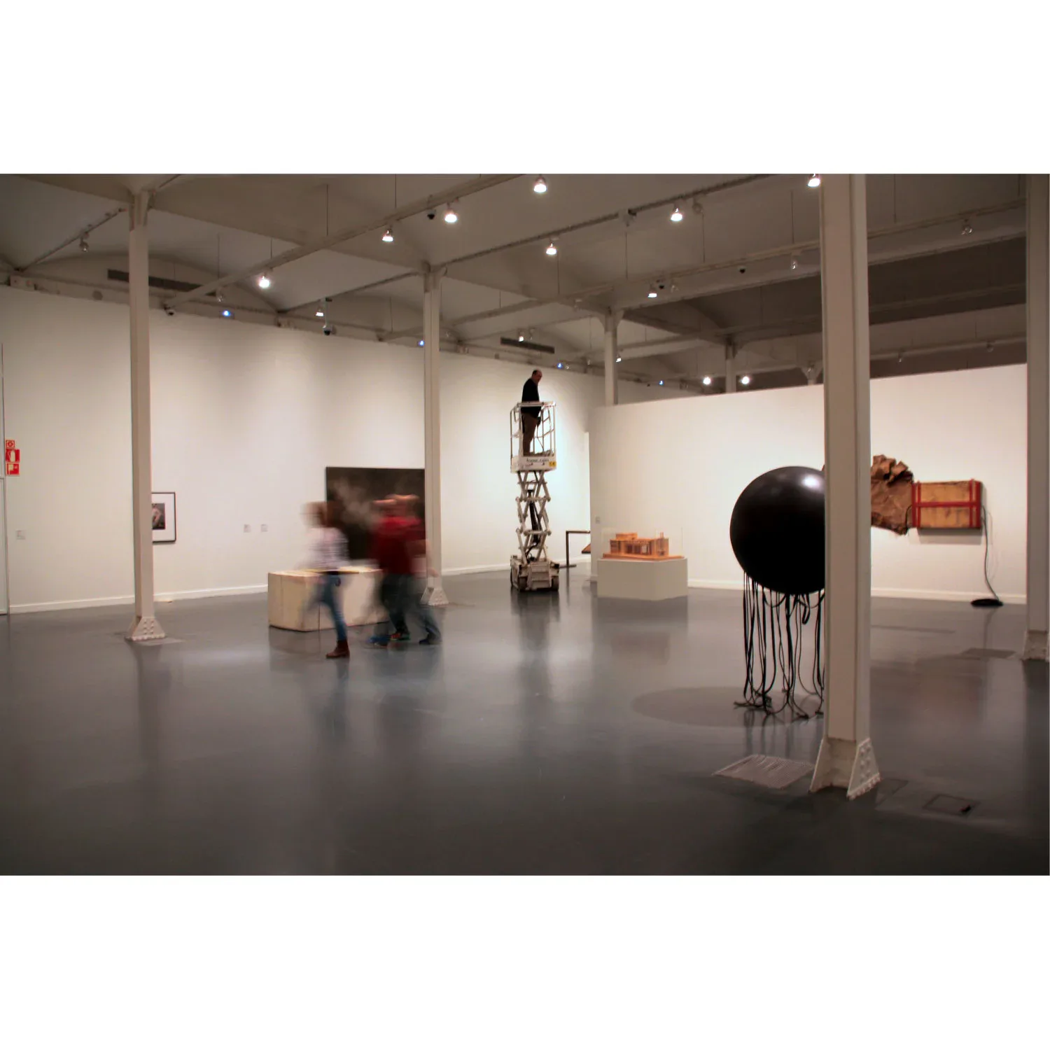 Minimalist gallery space with polished concrete floors, white walls, and structural columns displaying contemporary art installations including sculptural pieces and wall-mounted works by Angel Mombiedro (Armombiedro Studio).