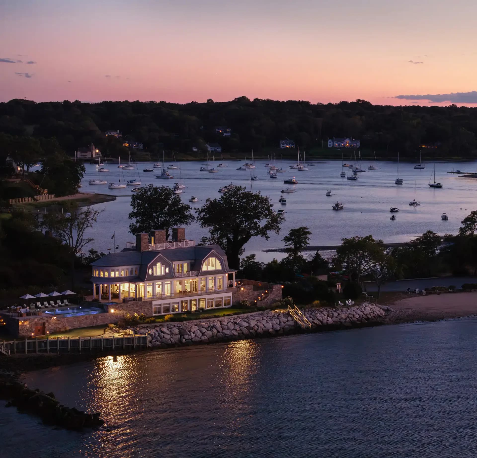 Waterfront residential estate with illuminated Victorian-style mansion at dusk, rocky shoreline, and sailboat-dotted harbor by Angela DeLouker.
