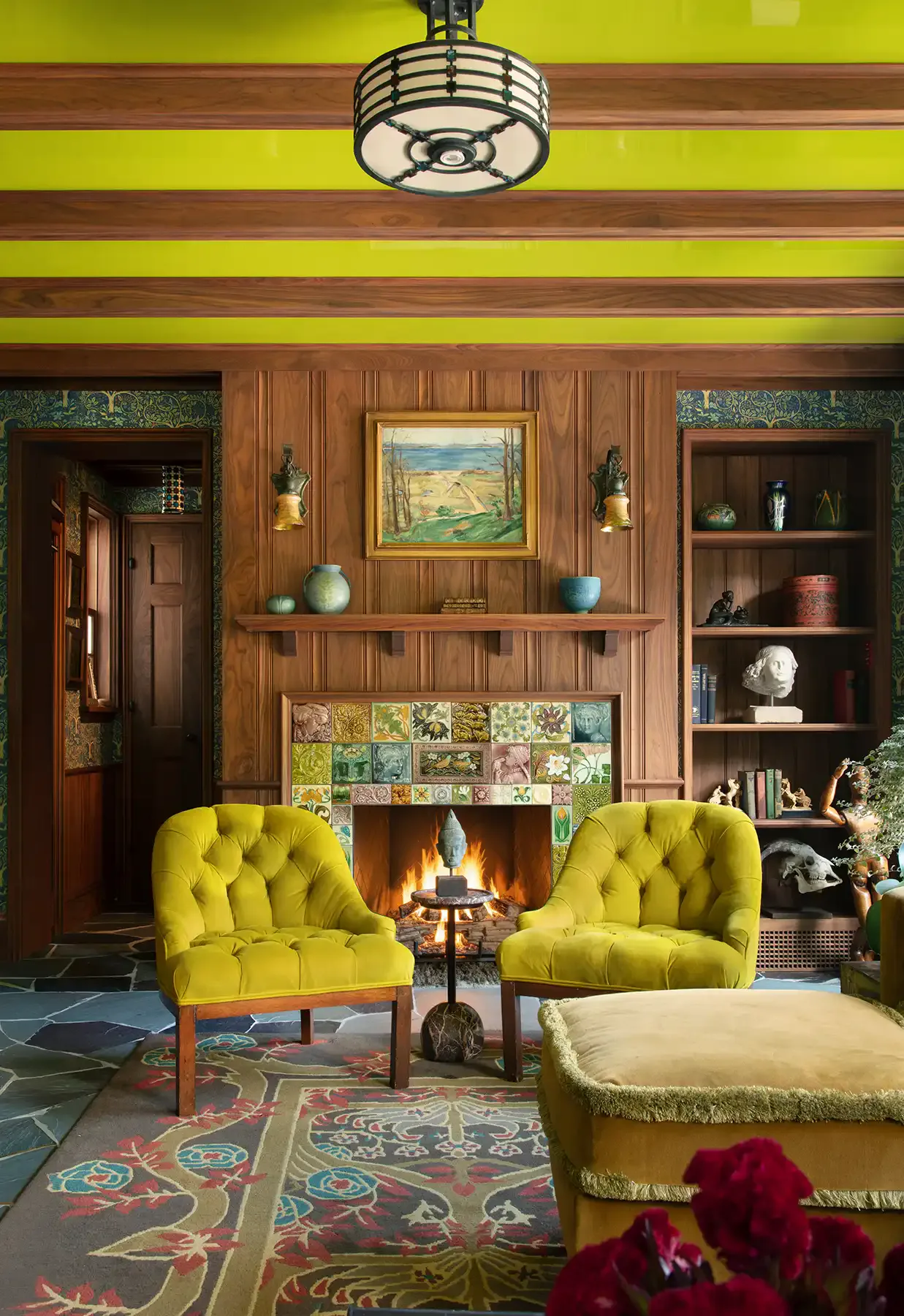 A striking living room with lime-green ceiling, wood-paneled walls, two chartreuse tufted chairs, fireplace with decorative tile surround, and patterned area rug by Angela DeLouker.