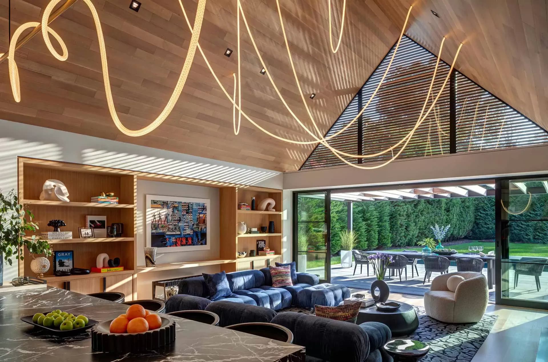 Modern living room with curved neon ceiling installations, navy sectional sofa, wood shelving, and expansive glass doors overlooking manicured grounds by Angela DeLouker.