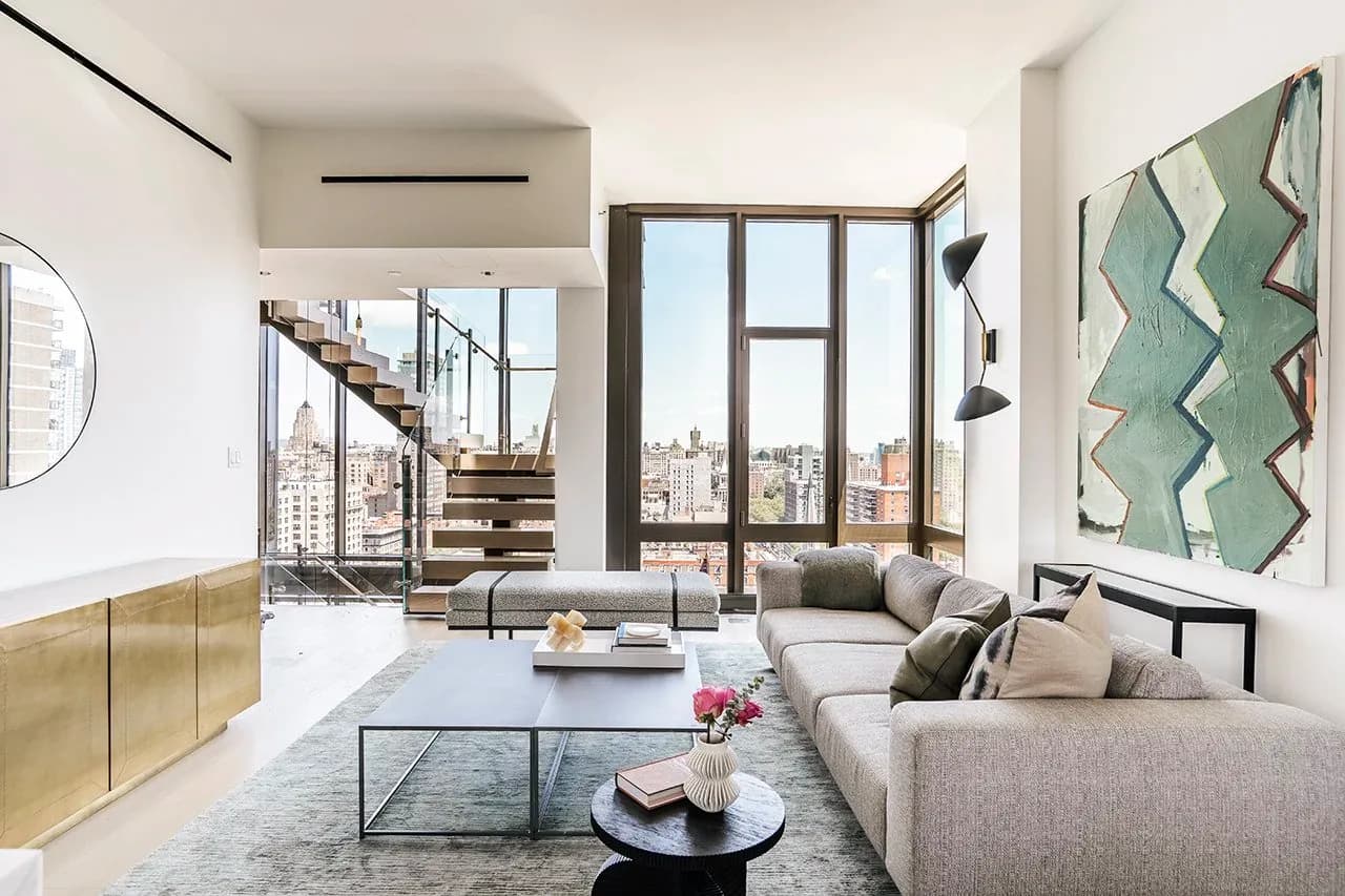 Modern loft living room with gray sofa, industrial staircase, floor-to-ceiling windows overlooking cityscape, geometric wall art, and minimalist furnishings by Angela Lee.
