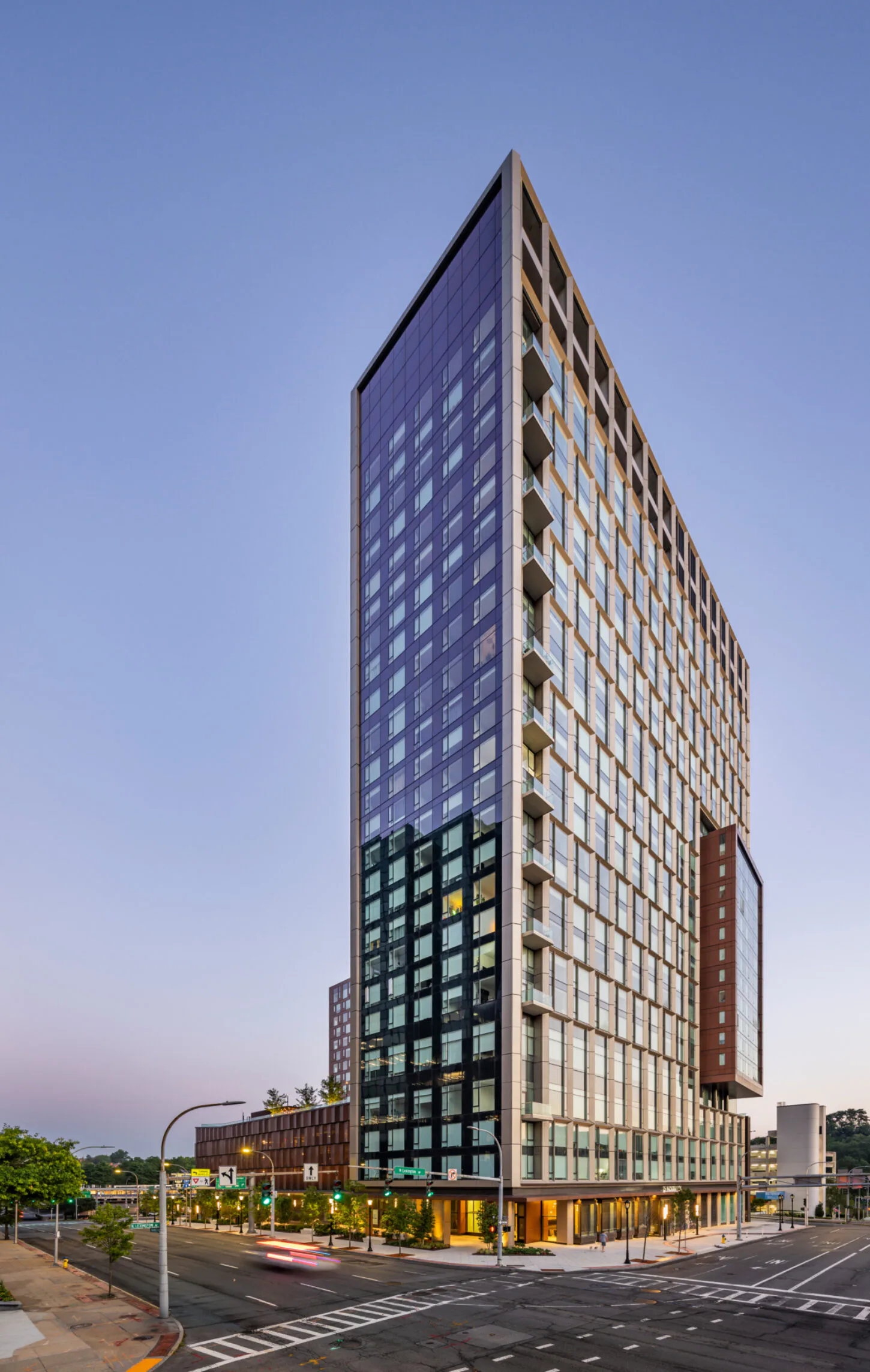 Tall modern mixed-use building with blue-tinted upper floors, cream and glass mid-section, and dark ground-level retail, photographed at dusk by Angela Liu.