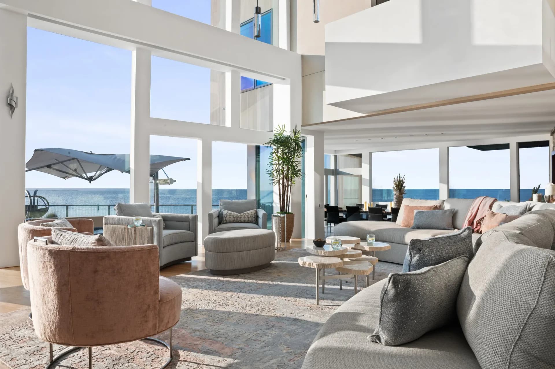 Luxurious coastal living room with floor-to-ceiling windows overlooking water and mountains, featuring neutral upholstered seating, natural materials, and clean modernist lines by Angelena Boeta.