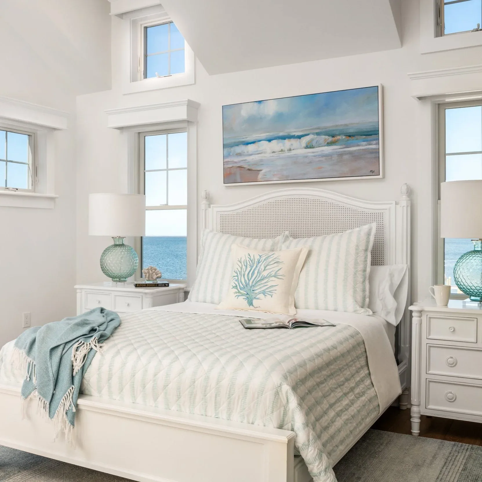 Bright coastal bedroom with white wainscot headboard, striped bedding, blue beach artwork, multiple windows, and seafoam accessories by Angelena Boeta.