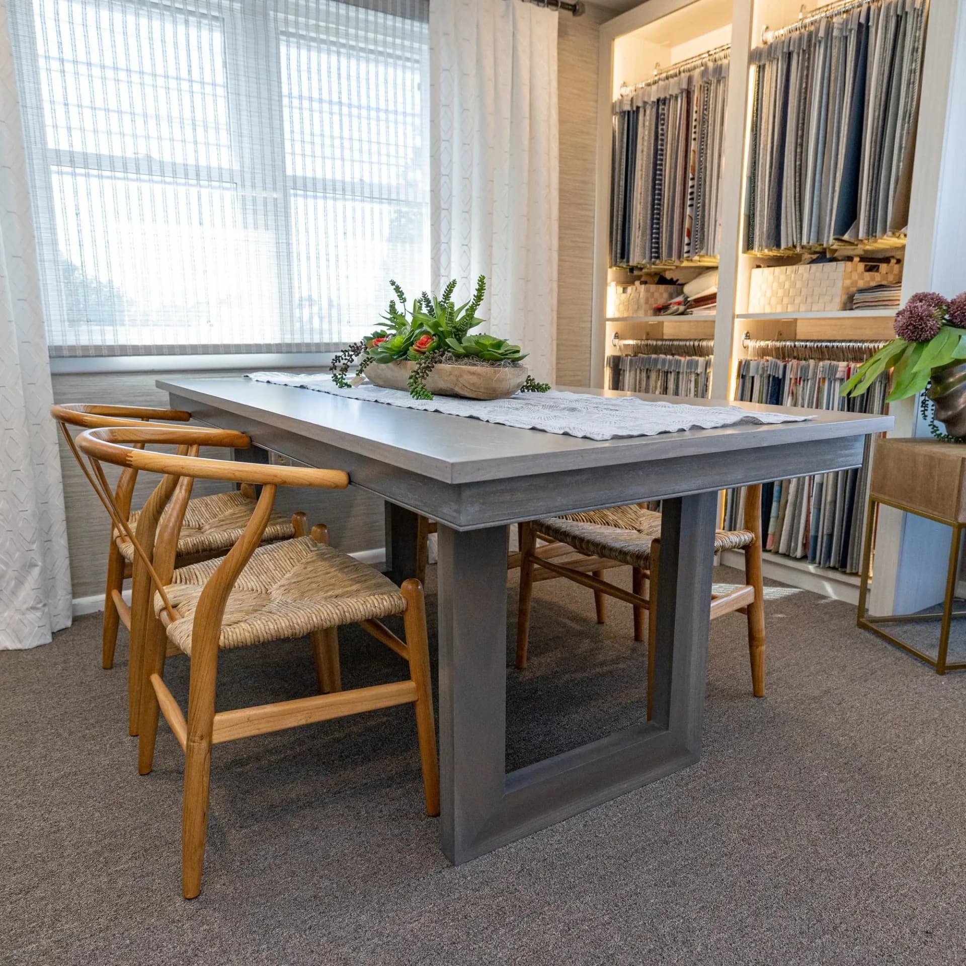 Modern showroom featuring a dark gray dining table with white marble top, wooden crossback chairs, and built-in shelving with fabric samples by Angelena Boeta.