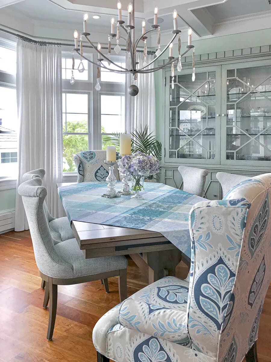 Elegant dining room with mint-green cabinetry, blue abstract table runner, upholstered chairs with geometric patterns, and a statement chandelier by Angelena Boeta.