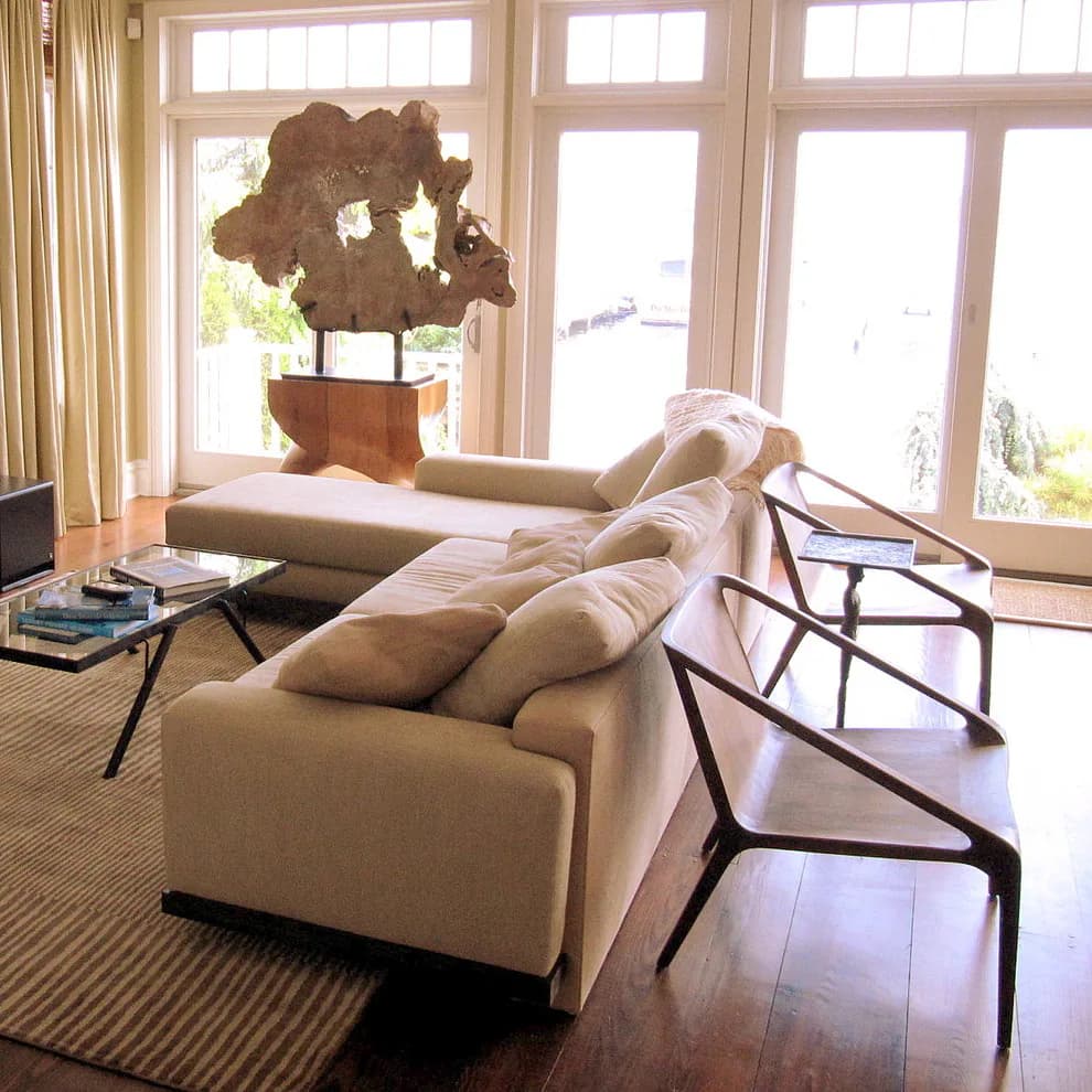 Bright contemporary living room with cream upholstered sectional, wooden dining chairs, dark hardwood floors, and potted sculptural plant by large transom windows, by Angelika Wrzesinska.