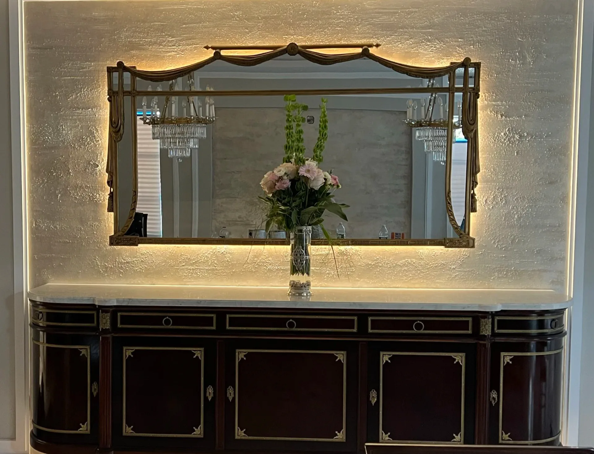 Luxurious bathroom vanity with illuminated mirror, dark wood cabinetry with brass detailing, and fresh floral arrangement by Ani Brieger.