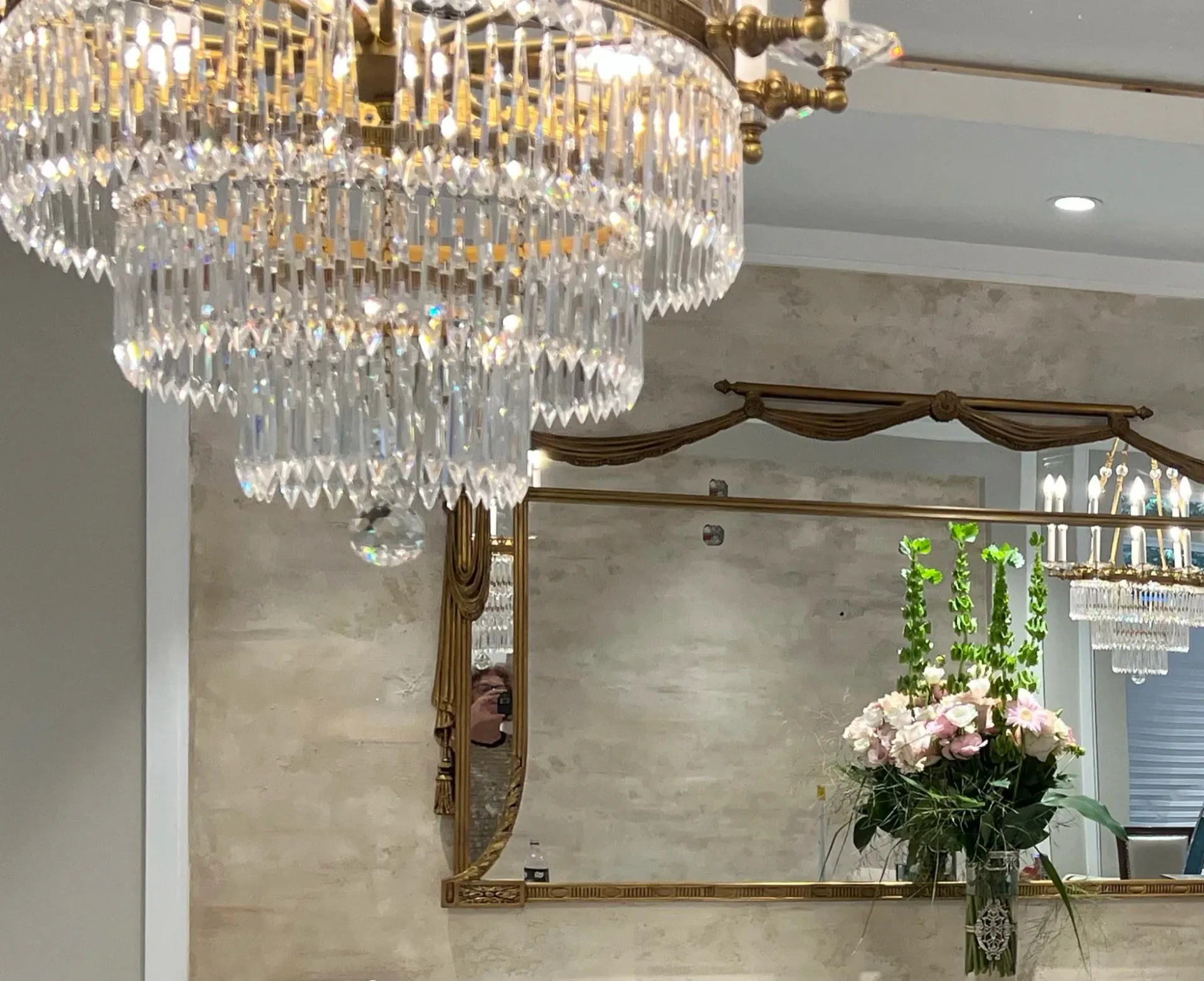 Luxurious bathroom featuring a multi-tiered crystal chandelier, brass-framed mirror, marble walls, and fresh floral arrangement by Ani Brieger.