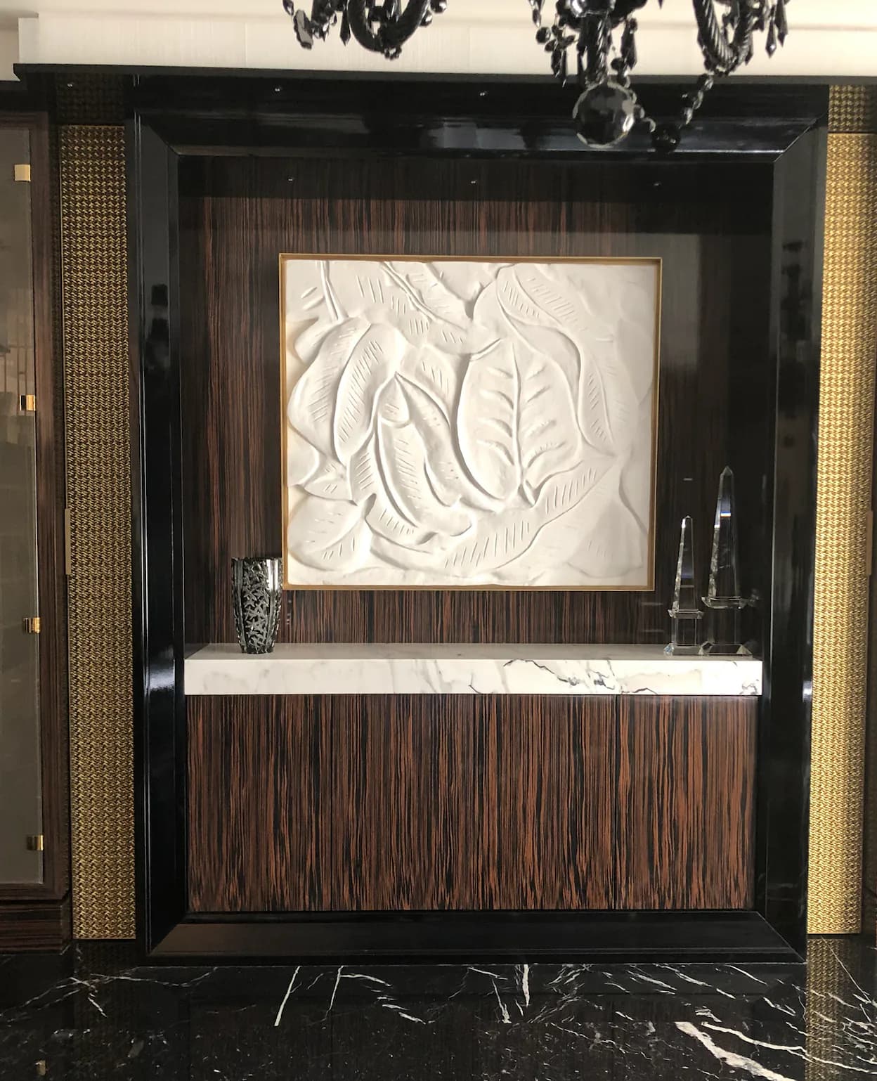 Modern spa reception desk with dark wood striped cabinetry, white marble countertop, and relief leaf artwork in gold frame by Ani Brieger.