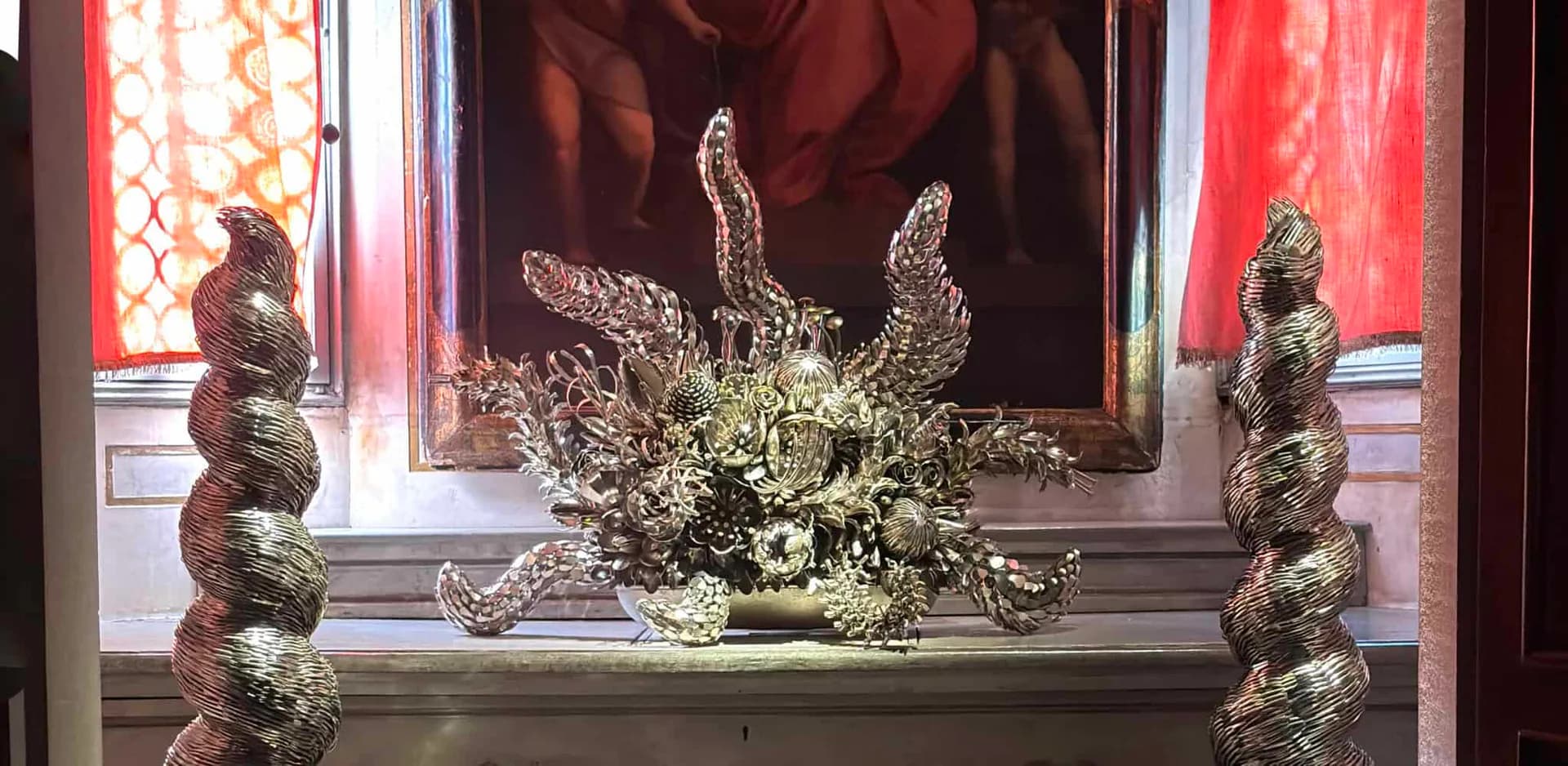 Ornamental display featuring two twisted bronze sculptural elements flanking a central floral arrangement with red silk backdrop, designed by Ann Carrington.
