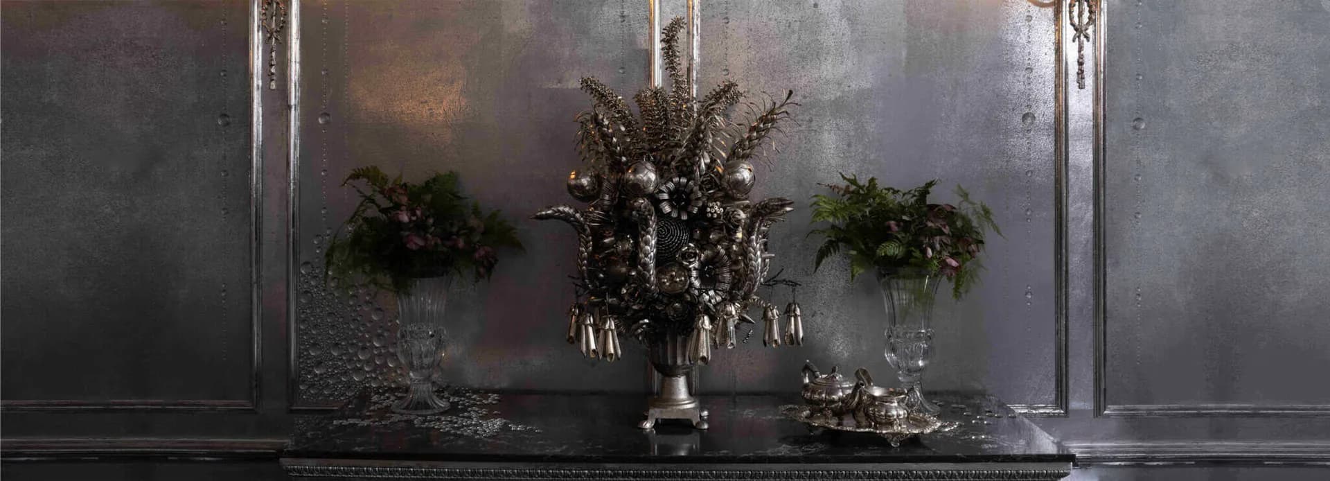 Dark moody mantelpiece with metallic wall panels, ornamental dried florals in urns, and candlesticks by Ann Carrington.