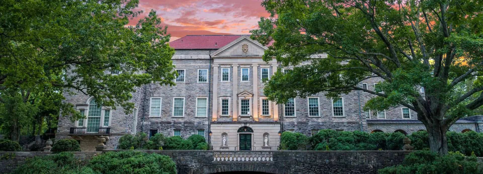 Elegant residential exterior at sunset featuring a grey Colonial-style home with blue shutters, manicured hedges, and mature trees framing the facade by Ann Carrington.