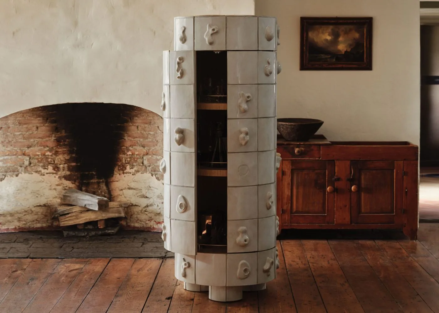 Rustic interior featuring a large brick fireplace, cylindrical white ceramic storage unit, wooden furniture, and terracotta floor by Anna Karlin.