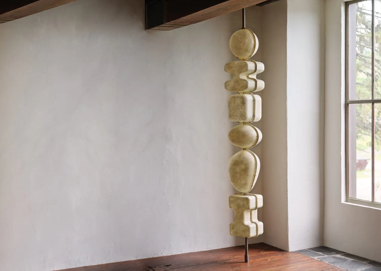 Minimalist interior installation featuring a vertical brass sculptural mobile with circular and rectangular elements suspended in a bright, white gallery space by Anna Karlin.