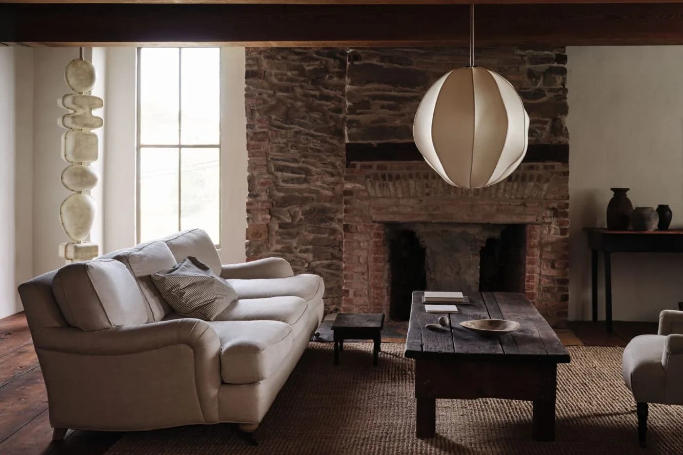 Contemporary living room with exposed brick fireplace, curved taupe sofa, black coffee table, pendant lighting, and warm earth-tone palette by Anna Karlin.