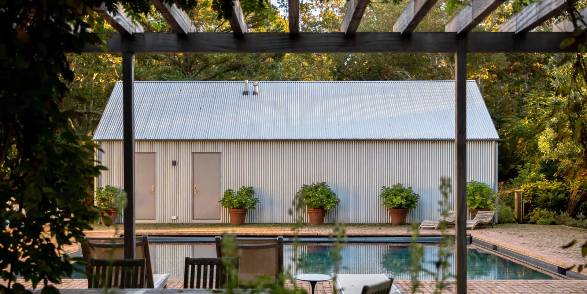 Modern poolside pavilion with steel pergola, corrugated metal backdrop, potted greenery, and lounge seating by Annabelle Selldorf.