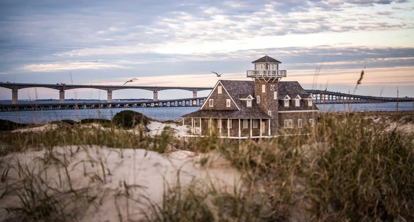 Victorian-era residential building with cupola situated in coastal dunes near a modern bridge at dusk, by Annette Veliz.