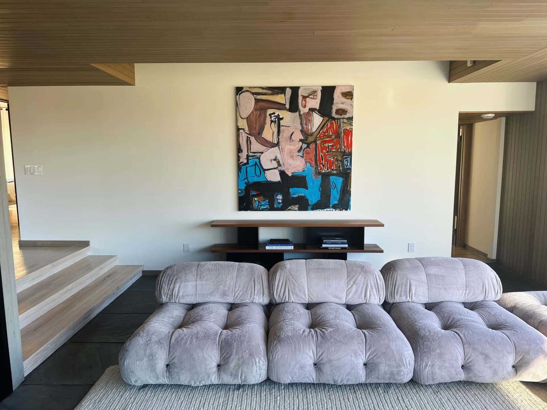 Modern bedroom interior featuring a sculptural puffy gray upholstered bed frame, floating black shelving with artwork above, and minimalist furnishings by Annie Nelson.