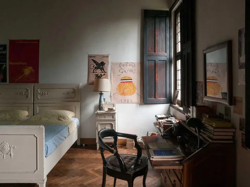 Bedroom interior with cream-colored bed, wooden desk, black chair, and artwork on walls; warm wood flooring and natural light by Annie Nelson.