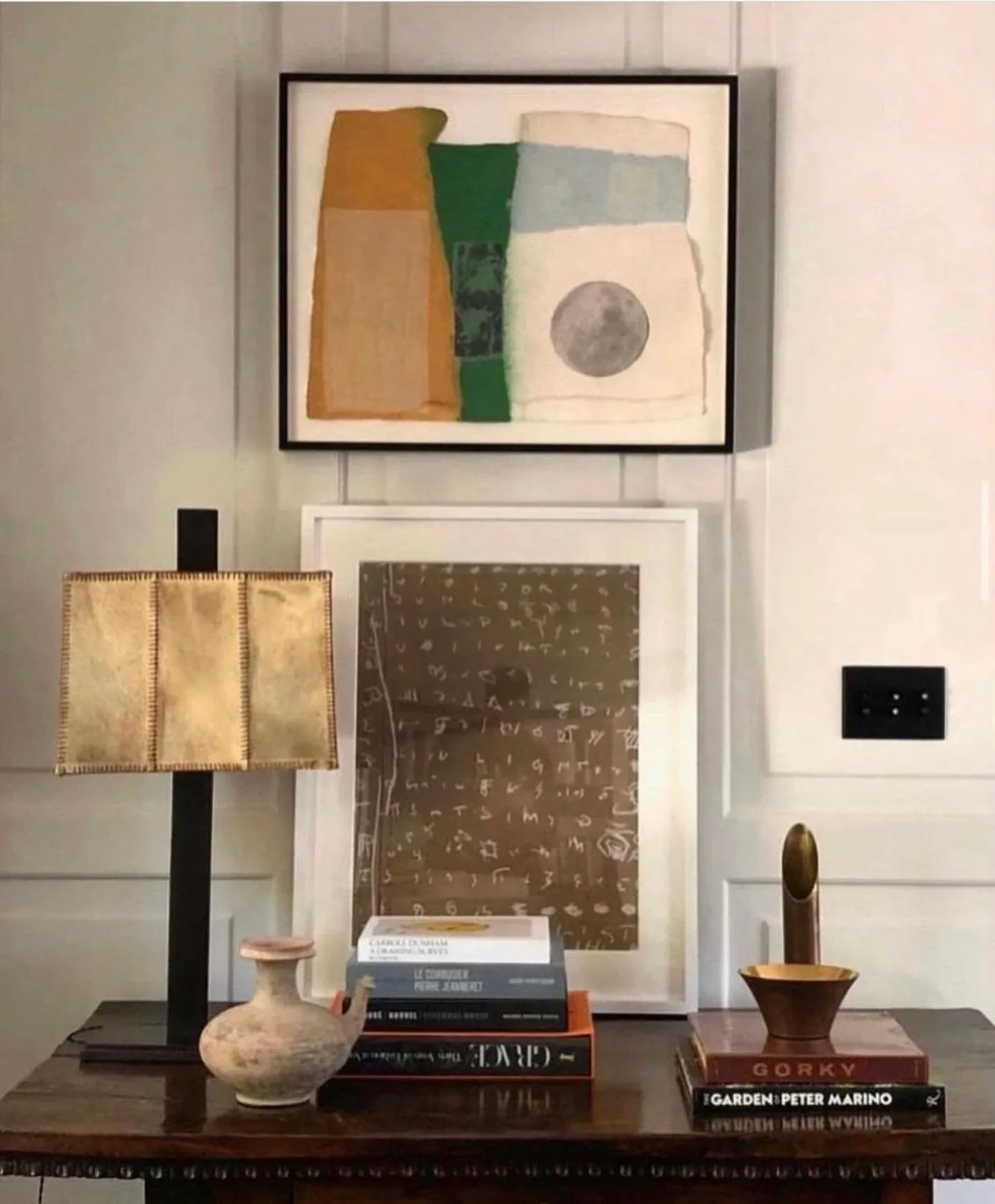 Styled console display with framed abstract artwork, decorative objects, books, and a table lamp arranged on dark wood furniture by Annie Nelson.