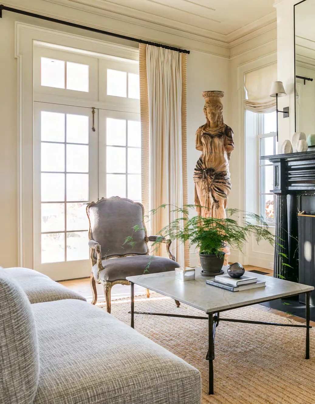A bright living room with cream walls, large French doors, upholstered seating, wooden sculpture, potted plant, and minimalist table designed by Antony Todd.