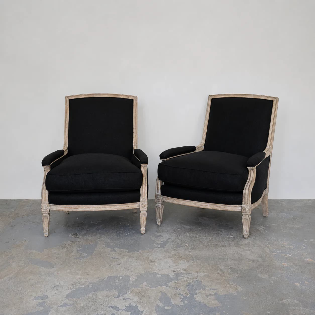 Two pale wooden Louis XVI-style armchairs upholstered in black fabric with turned legs, positioned in a minimalist gray concrete space by Antony Todd.