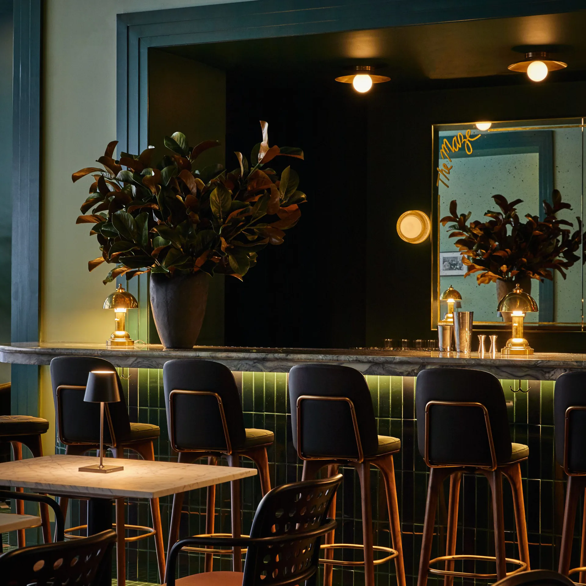 Modern bar interior with dark green banquette seating, black chairs with brass accents, wooden tables, potted plants, and warm ambient lighting by Aria Jahanshahi.