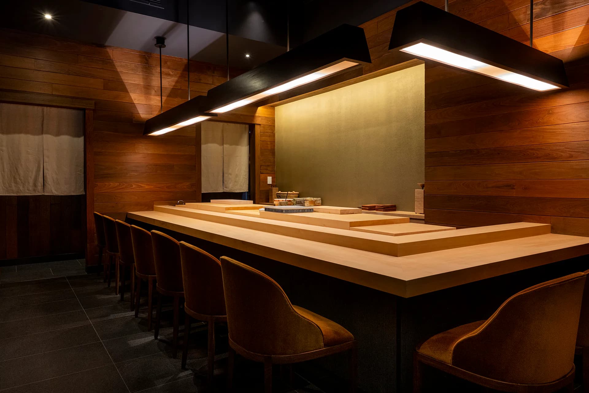 Modern sushi bar with continuous light wood counter, warm ambient lighting, dark wood walls, and curved leather seating by Aria Jahanshahi.