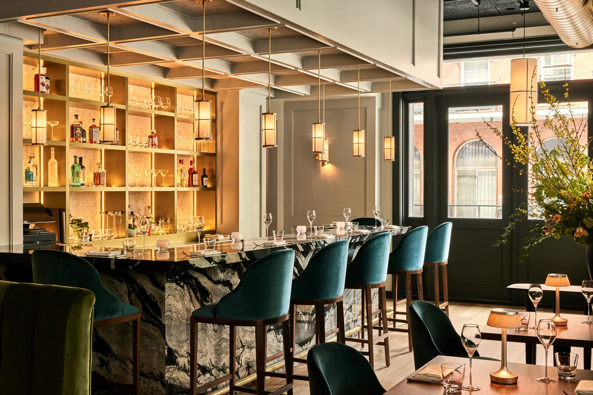 Modern bar and dining space with teal velvet seating, warm wood shelving, pendant lighting, and black architectural elements by Aria Jahanshahi.
