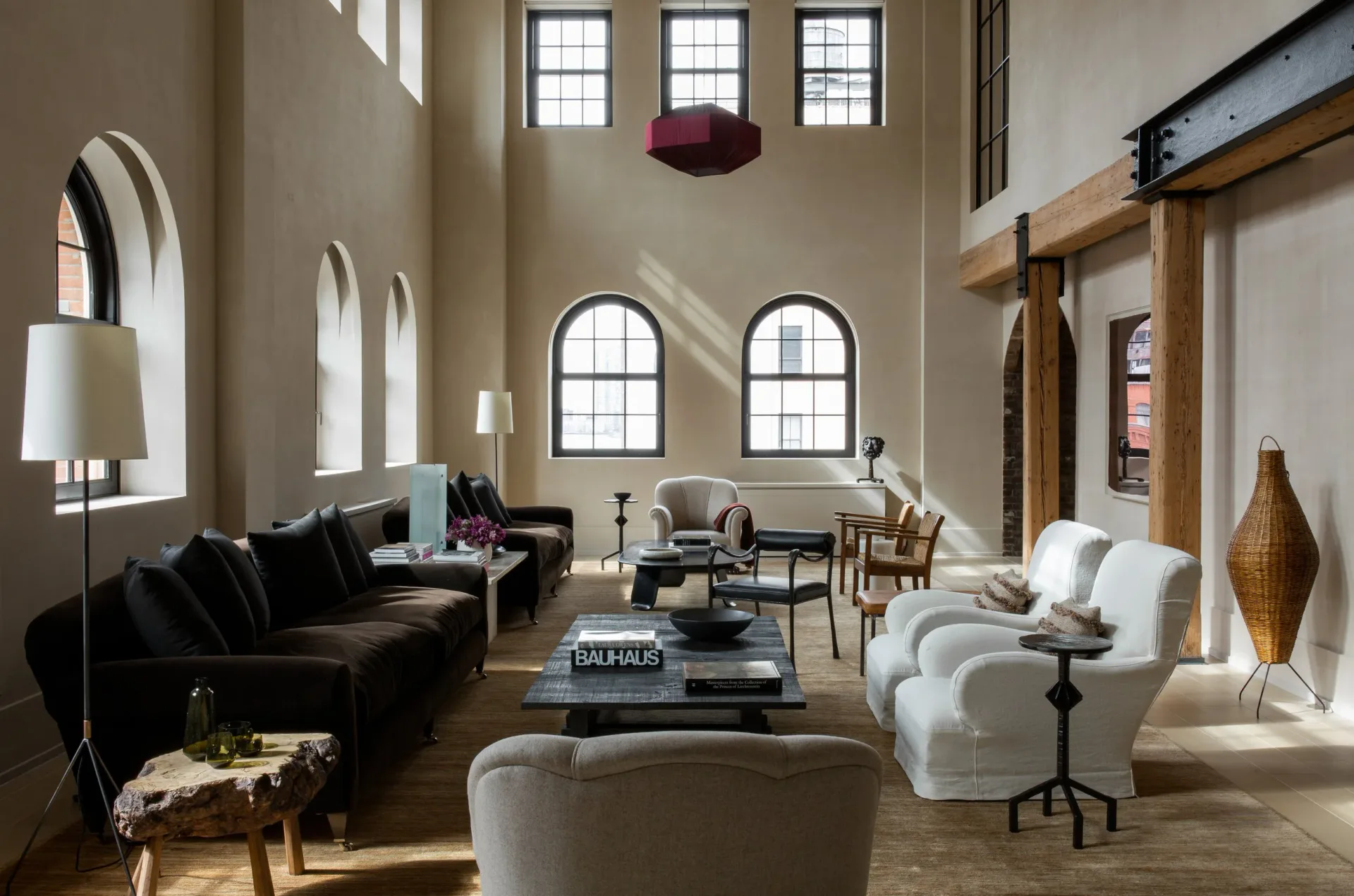 A spacious loft-style living room with soaring ceilings, arched windows, black and cream furnishings, wooden beams, and natural light, designed by Ariel Ashe.