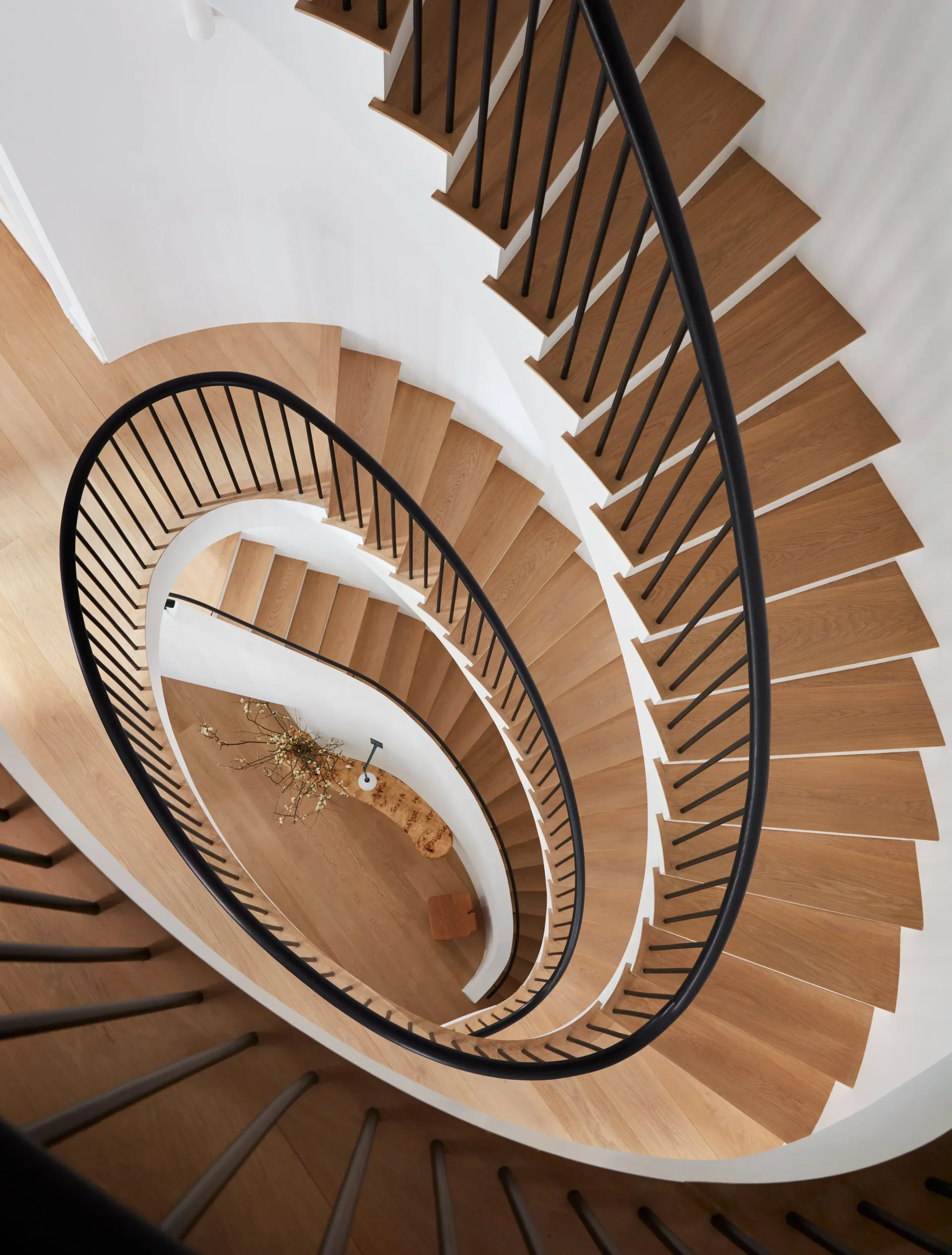 Overhead view of a spiral staircase with natural wood steps and black metal railings descending through white walls by Ariel Ashe.