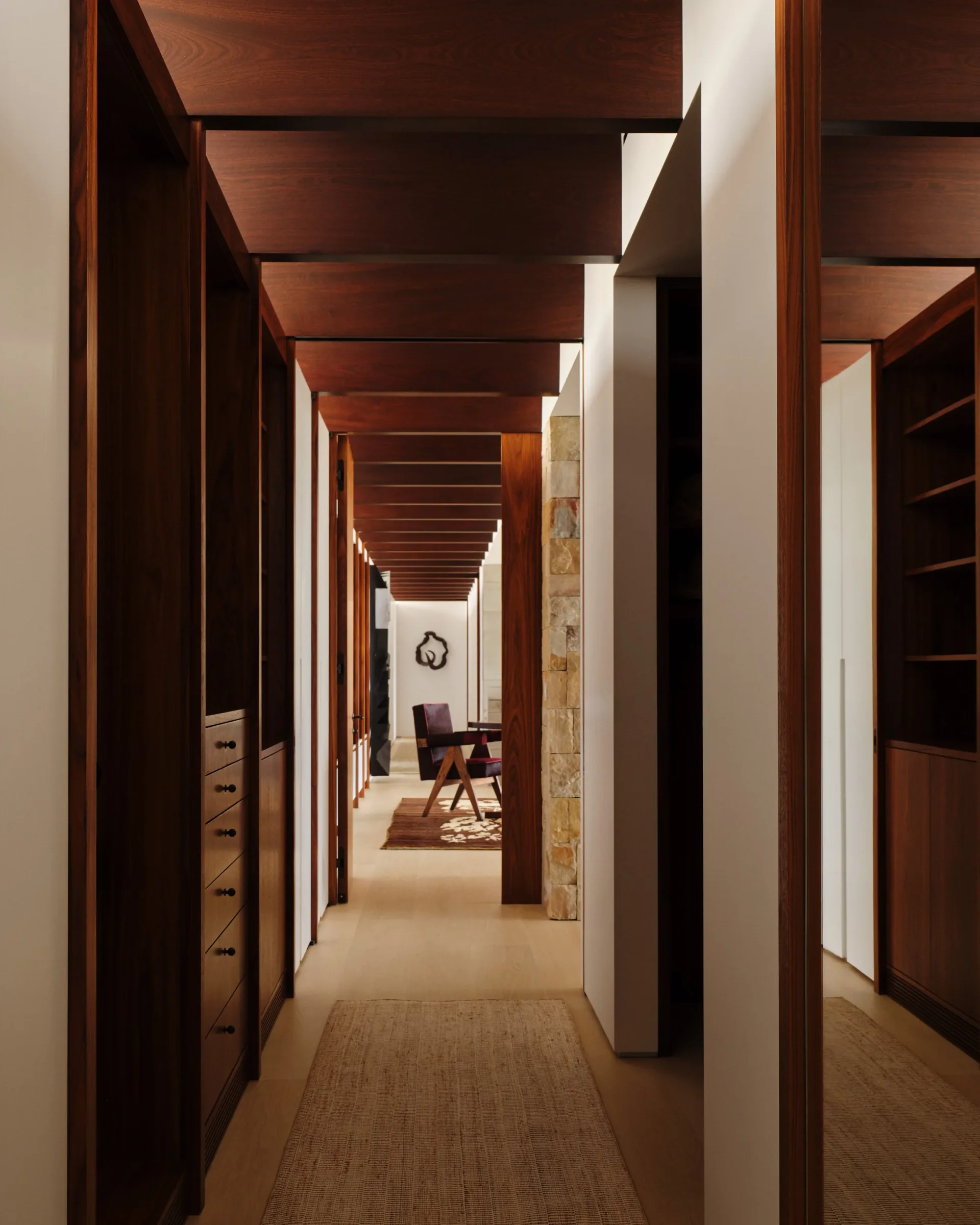 Long wooden corridor with dark timber beams and white walls leading to a lounge area, featuring natural light and warm materiality by Ariel Ashe.