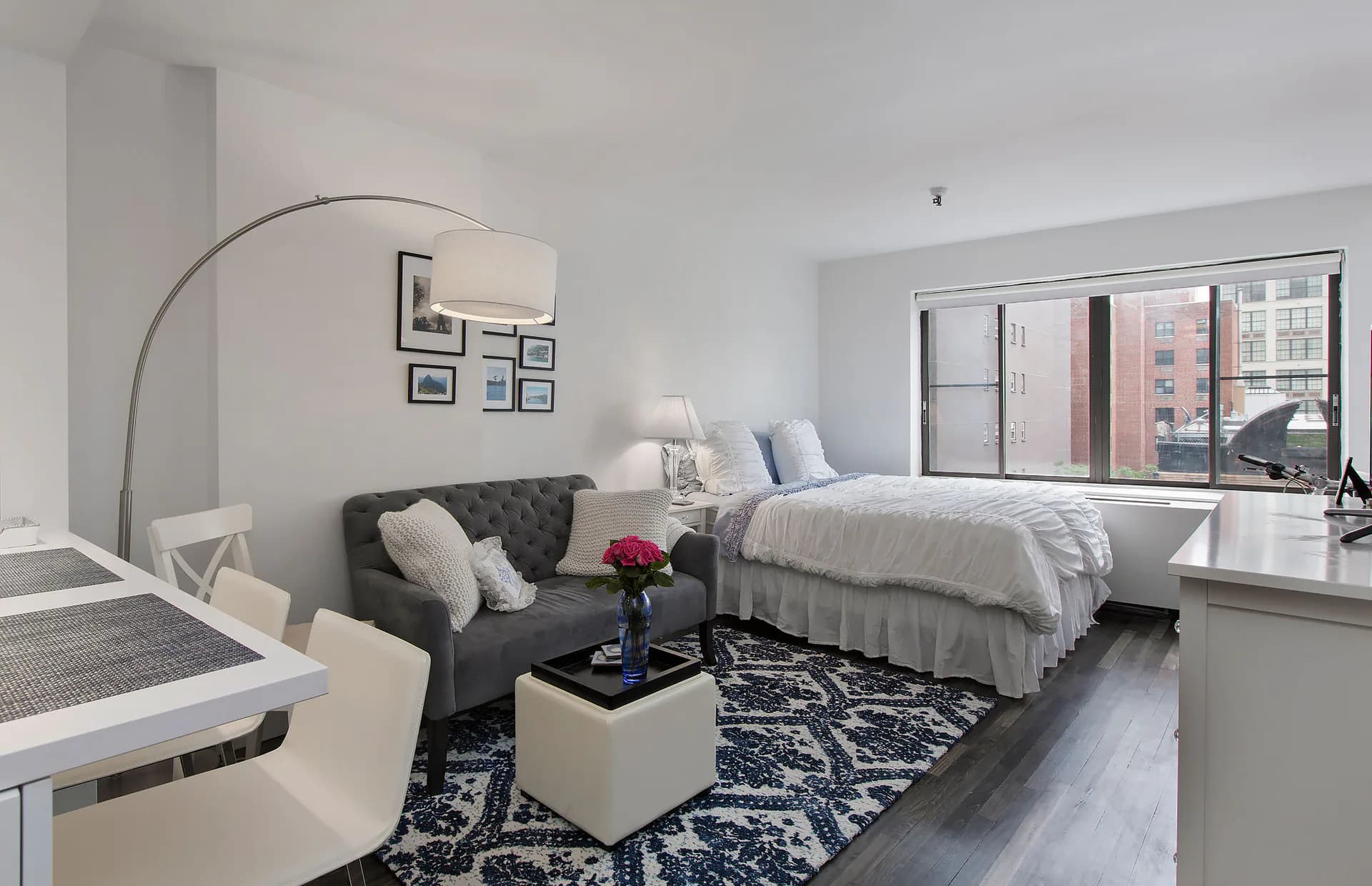 Modern studio apartment with grey upholstered seating, queen bed with grey bedding, arc floor lamp, dark patterned area rug, and floor-to-ceiling windows overlooking city skyline by Arlene Avidan.