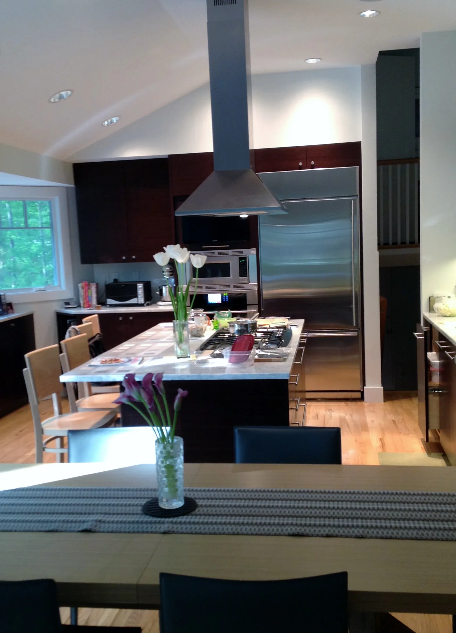 Modern kitchen and dining area featuring dark cabinetry, stainless steel appliances, island counter with white countertop, pendant hood vent, and natural wood flooring designed by Arlene Avidan.
