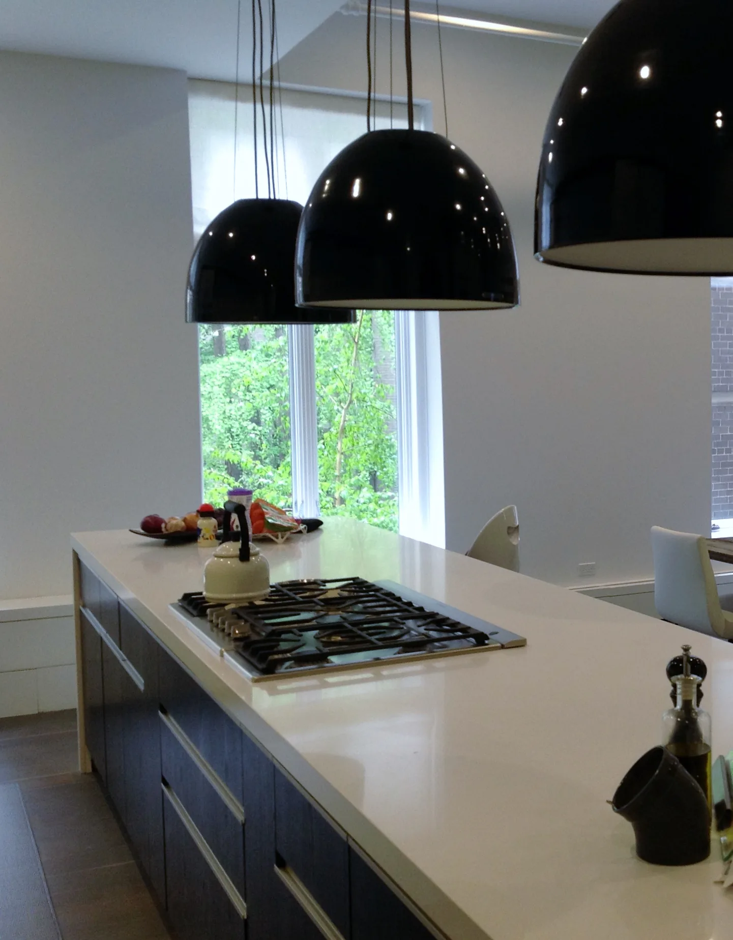 Modern kitchen with white quartz countertops, gas cooktop, and three black dome pendant lights suspended above an island, featuring floor-to-ceiling windows by Arlene Avidan.